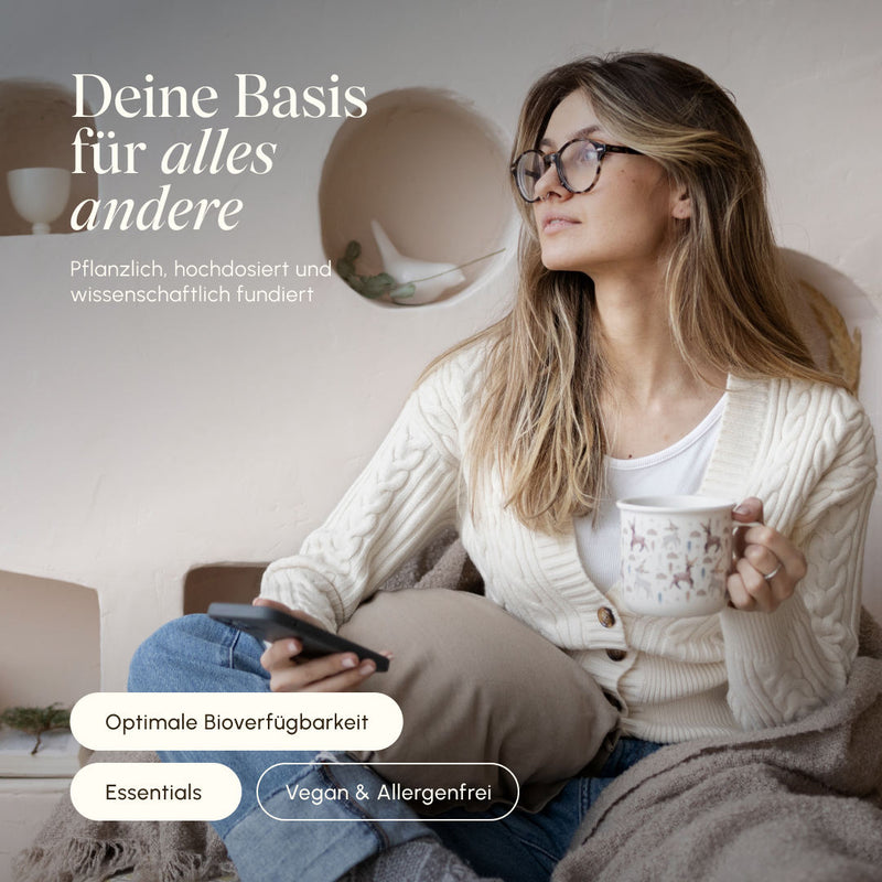Frau mit Brille sitzt entspannt auf dem Sofa mit einer Tasse Tee und Smartphone. Text: „Deine Basis für alles andere – pflanzlich, hochdosiert und wissenschaftlich fundiert". Badges: Optimale Bioverfügbarkeit, Essentials, Vegan & Allergenfrei