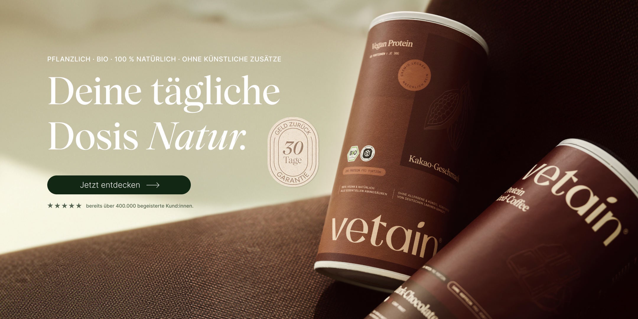 Vetain Vegan Protein Hero-Banner mit zwei Dosen (Kakao-Geschmack und Iced-Coffee) auf braunem Stoff. Headline „Deine tägliche Dosis Natur." mit Button „Jetzt entdecken", 30-Tage-Geld-zurück-Siegel und Hinweis „bereits über 400.000 begeisterte Kund:innen".
