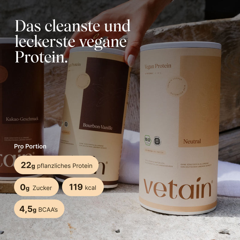 Drei vetain Vegan Protein Dosen (Kakao-Geschmack, Bourbon-Vanille, Neutral) auf Steinoberfläche, eine Hand greift nach der mittleren Dose. Im Vordergrund die Neutral-Dose. Text: „Das cleanste und leckerste vegane Protein." Pro Portion: 22g pflanzliches Protein, 0g Zucker, 119 kcal, 4,5g BCAAs.