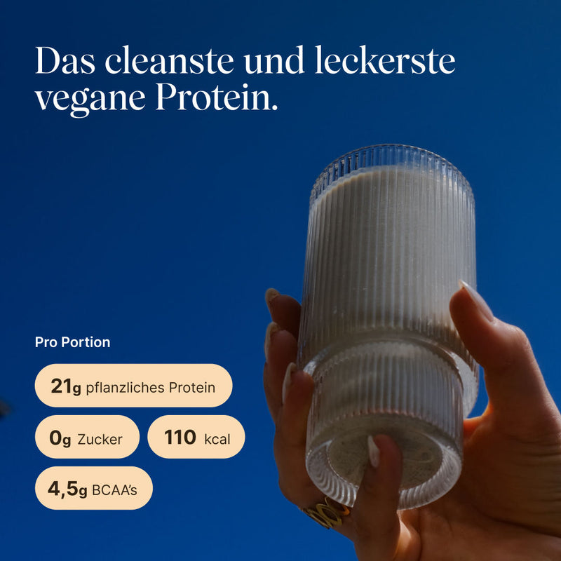 Hand hält ein geripptes Glas mit hellem Zimt-Proteinshake vor blauem Himmel. Text: „Das cleanste und leckerste vegane Protein." Pro Portion: 21g pflanzliches Protein, 0g Zucker, 110 kcal, 4,5g BCAAs.