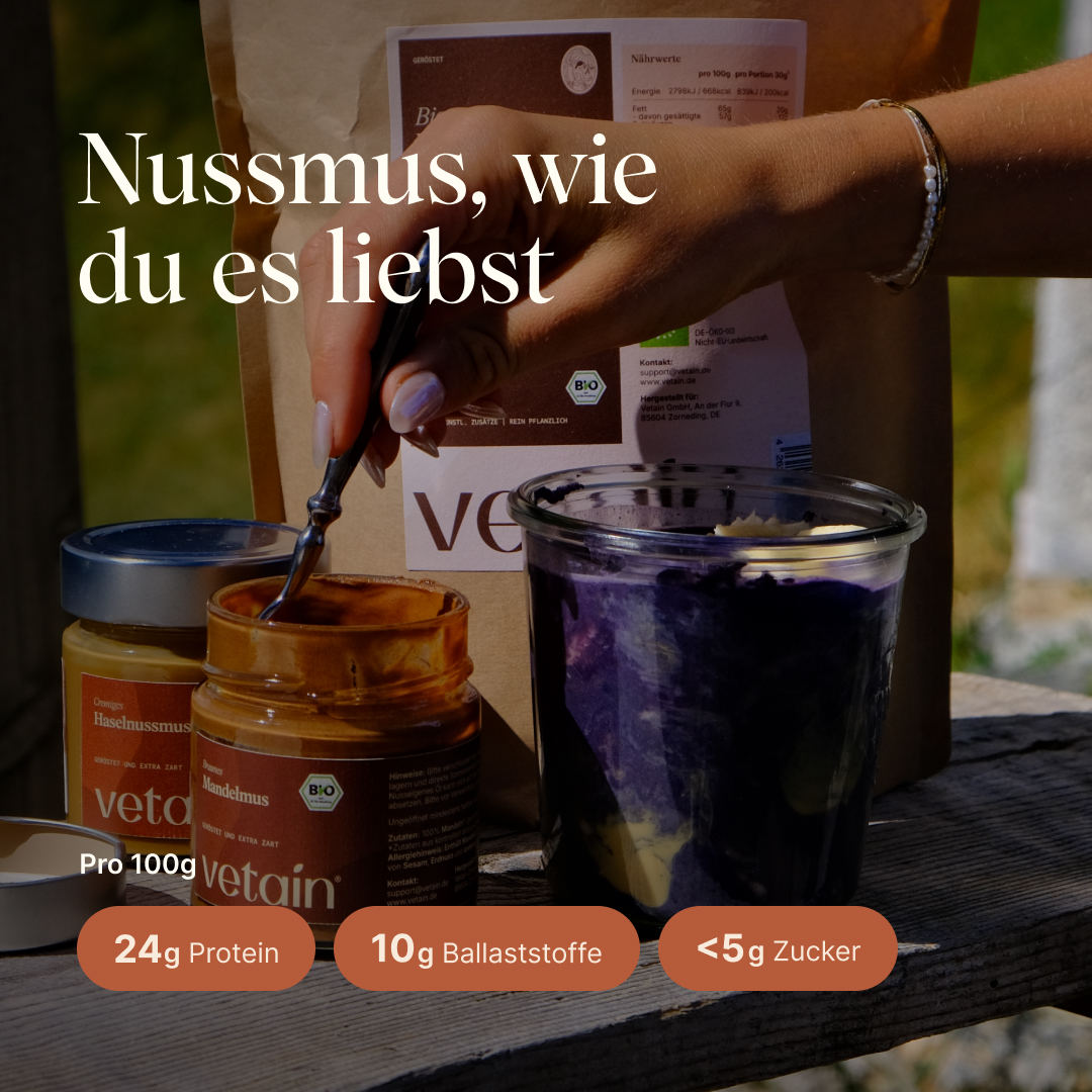 Nussmusglas mit Löffel darin, daneben ein Dessertglas mit Porridge darin. Ein Nussmus zum lieben mit 24g Protein, 10g Ballaststoffen und unter 5g Zucker pro 100g0