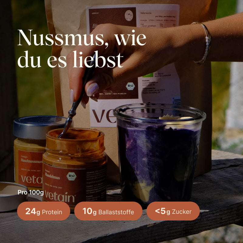 Nussmusglas mit Löffel darin, daneben ein Dessertglas mit Porridge darin. Ein Nussmus zum lieben mit 24g Protein, 10g Ballaststoffen und unter 5g Zucker pro 100g0